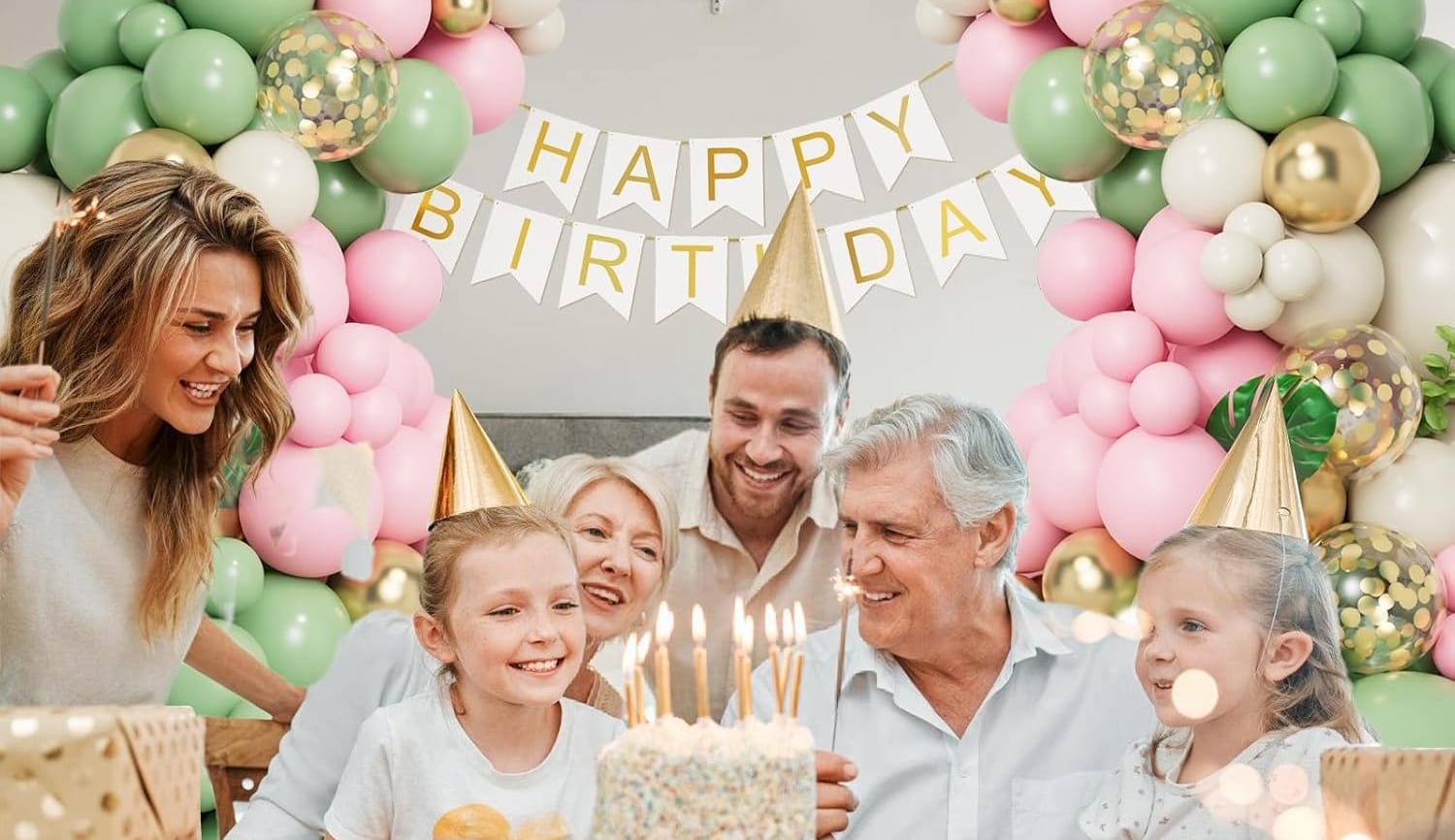 A family celebrating a birthday with a cake in front of a pink, green and gold balloon garland